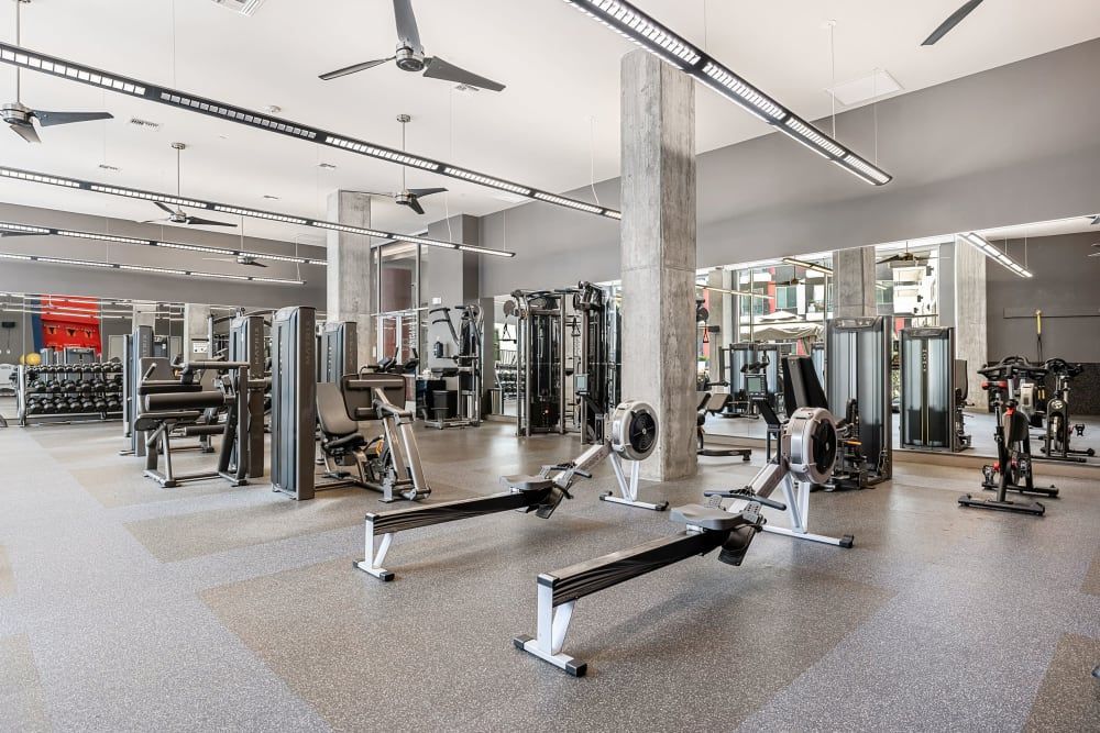 A modern and spacious gym with various workout machines and equipment, including weight benches, leg press, rowing machines, and racks of dumbbells, set on a speckled floor with structural columns and large mirrors. The gym is well-lit by natural light and overhead linear lights, with ceiling fans to provide airflow at Marq on Burnet in Austin, TX.