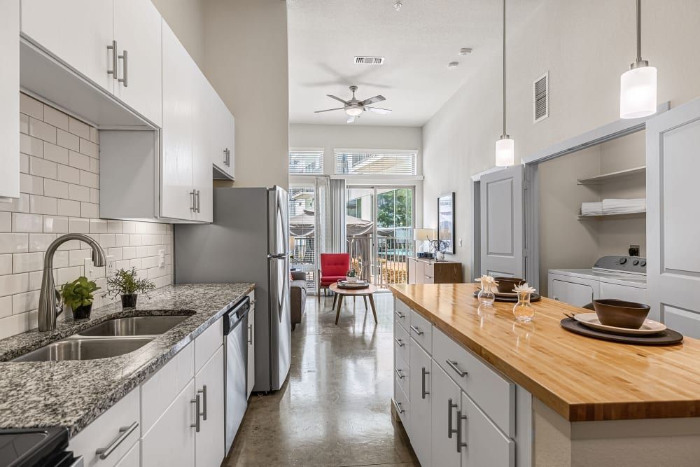 Modern kitchen interior with white cabinetry, stainless steel appliances, and a wooden countertop island. The open concept layout reveals a dining area with a red chair in the background, leading to a balcony with natural light streaming in at Marq on Burnet in Austin, TX.
