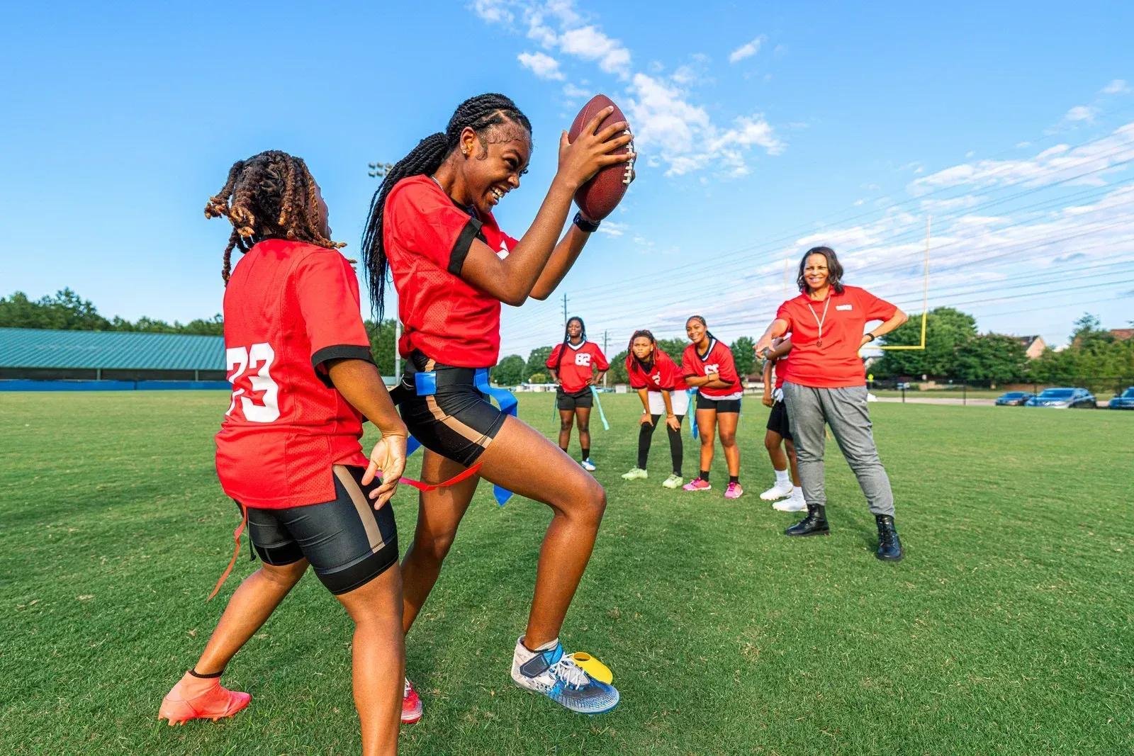 Women playing flag football on a grassy field. One catches the ball while another defends. Others watch. Red and black uniforms.