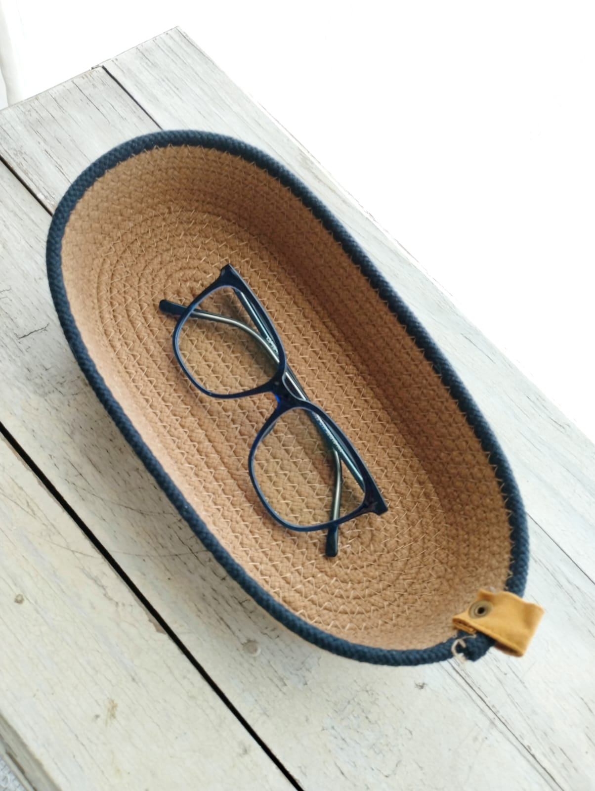 Gafas negras en una bandeja ovalada tejida de color tostado sobre una superficie de madera blanca.