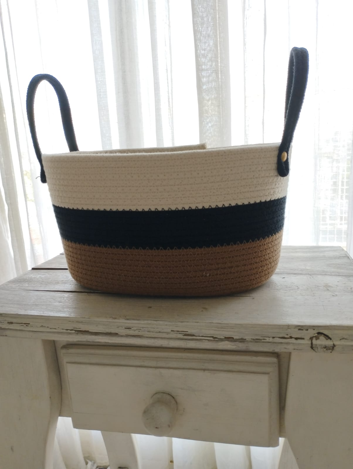 Cesta tejida bicolor con asas negras sobre una mesa blanca envejecida junto a una ventana.