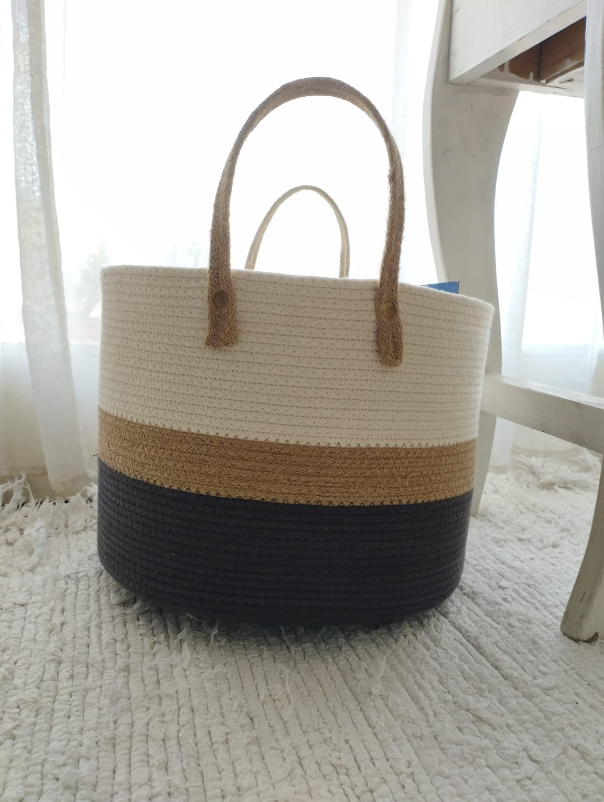 Bolso de tela con rayas beige y negras sobre una alfombra cerca de una silla y una ventana.