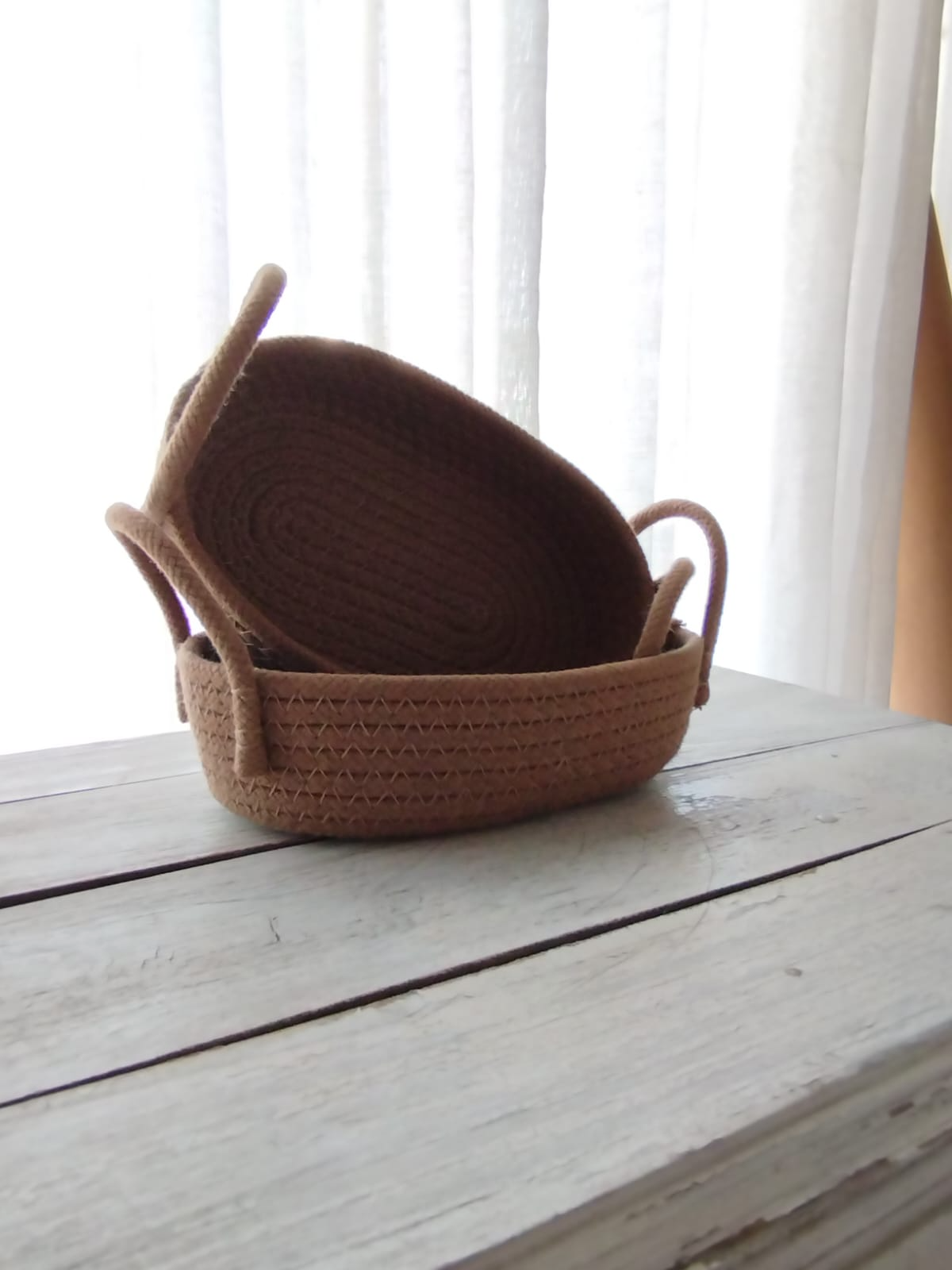 Cesta tejida con un asa en relieve sobre una mesa de madera blanca junto a una ventana luminosa.