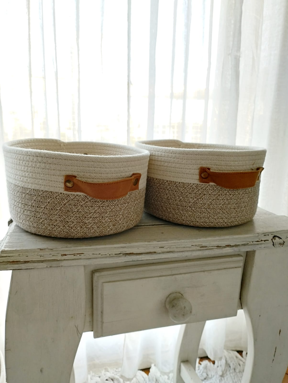 Dos cestas de almacenamiento tejidas con asas color canela sobre una mesa blanca desgastada junto a una ventana luminosa.