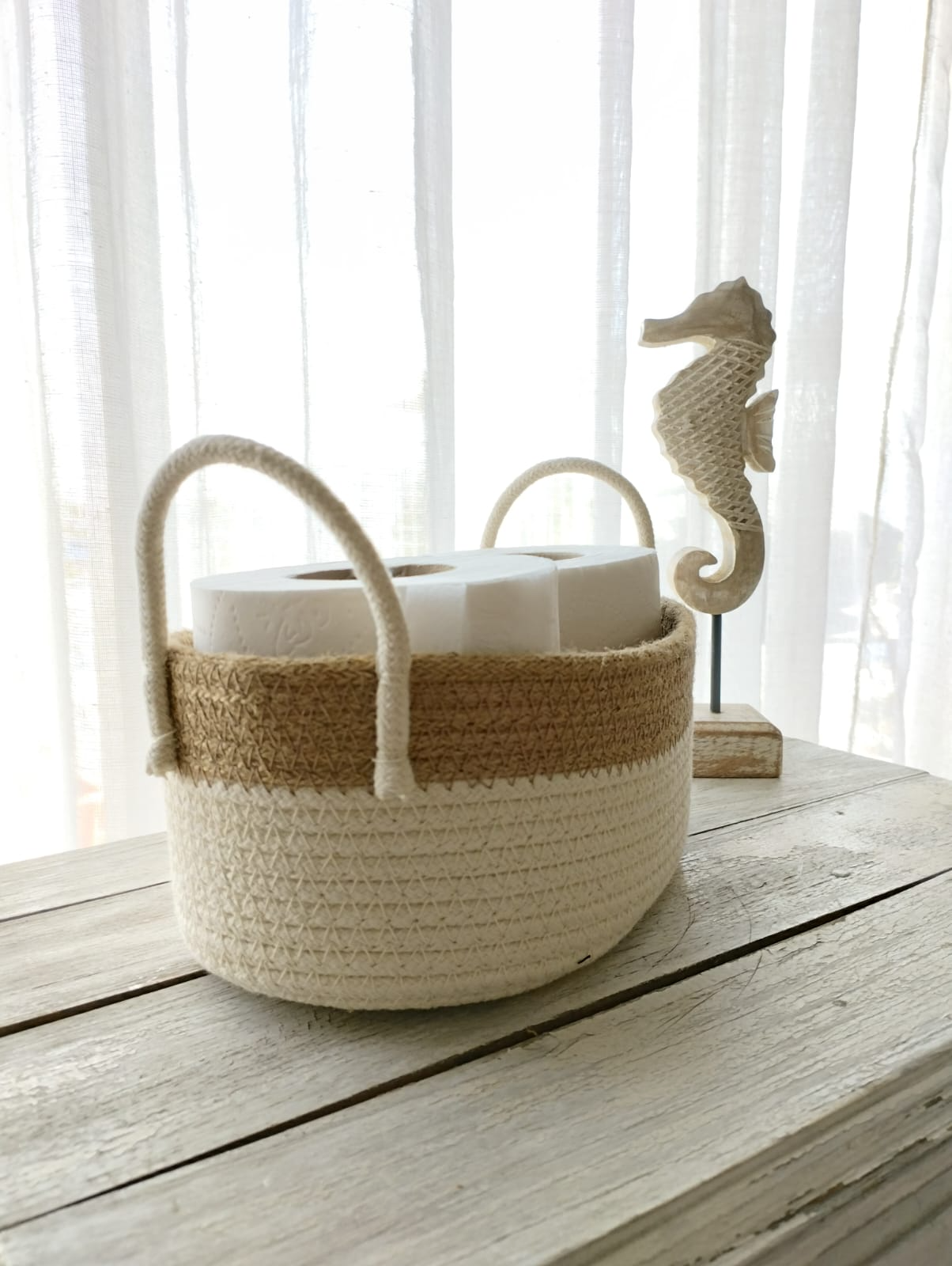 Cesta tejida bicolor con asas de cuerda y adorno de caballito de mar sobre una mesa rústica blanca.