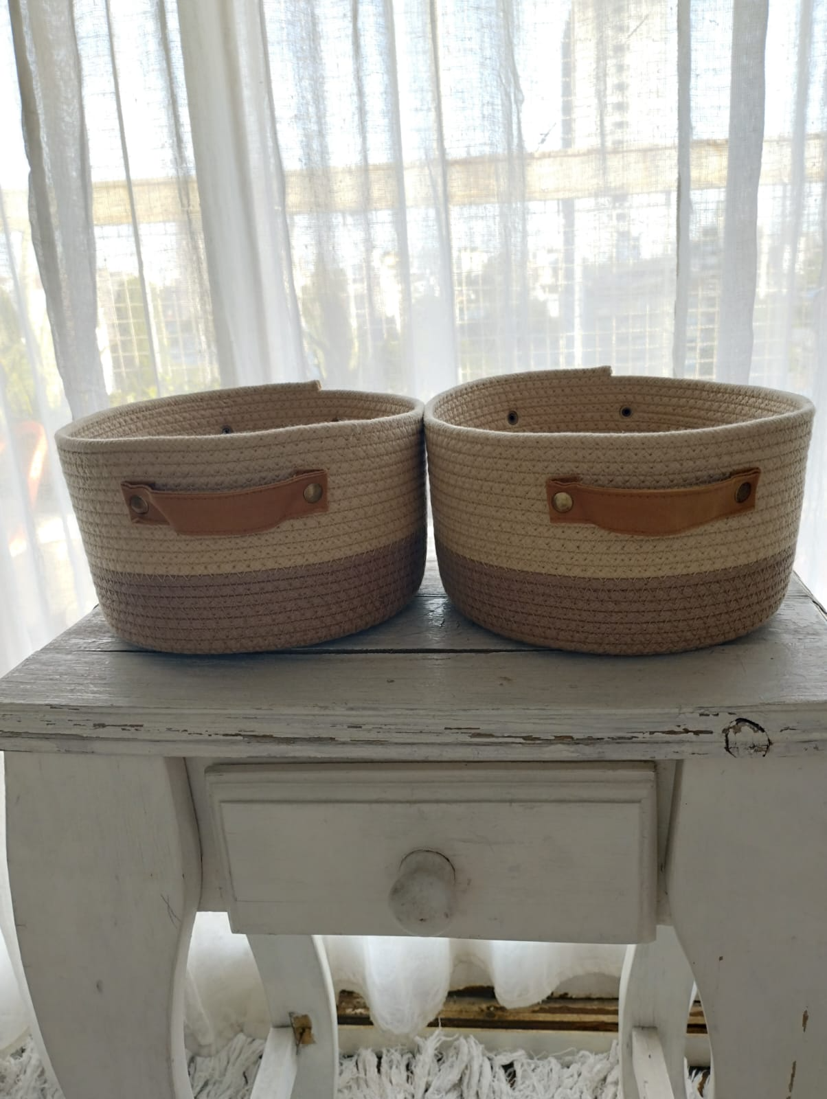 Cesta de mimbre tejida con tapa, de color tostado y marrón, colocada sobre una mesa de madera blanca.