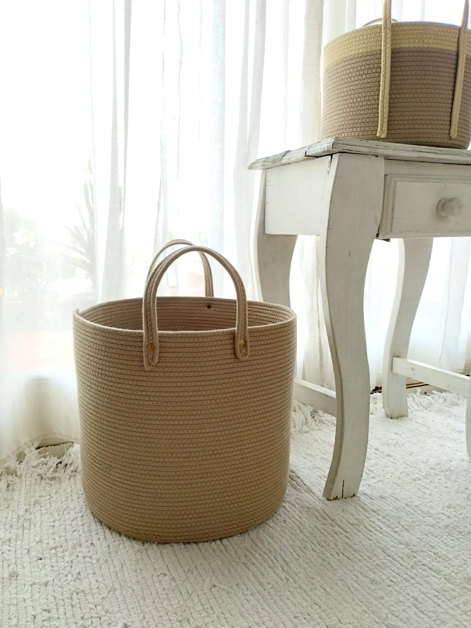 Dos cestas de mimbre tejidas en una habitación de color blanco brillante, junto a una mesa blanca de aspecto envejecido.
