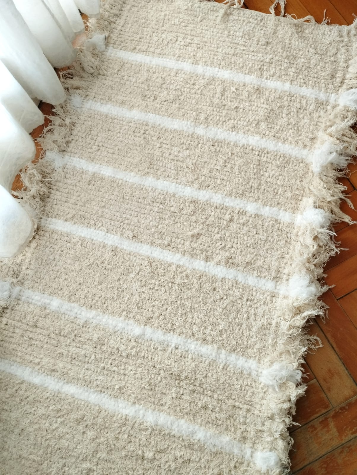 Alfombra blanca tejida con rayas marrones y bordes con flecos sobre un suelo de madera.
