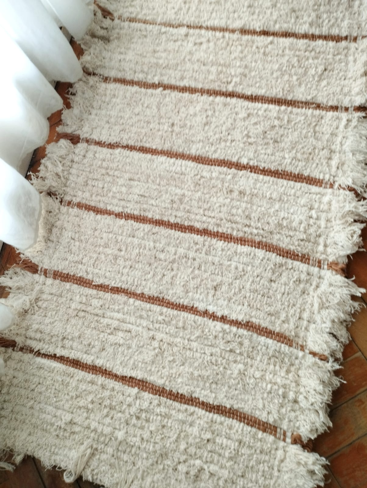 Alfombra blanca tejida con rayas marrones y bordes con flecos sobre un suelo de madera.