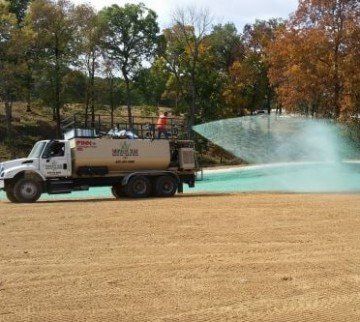 A truck spraying green liquid on a brown field. Trees and an overcast sky are in the background.