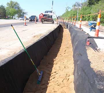 Road construction site with a long trench lined with black fabric, filled with sand. A street runs along the background.