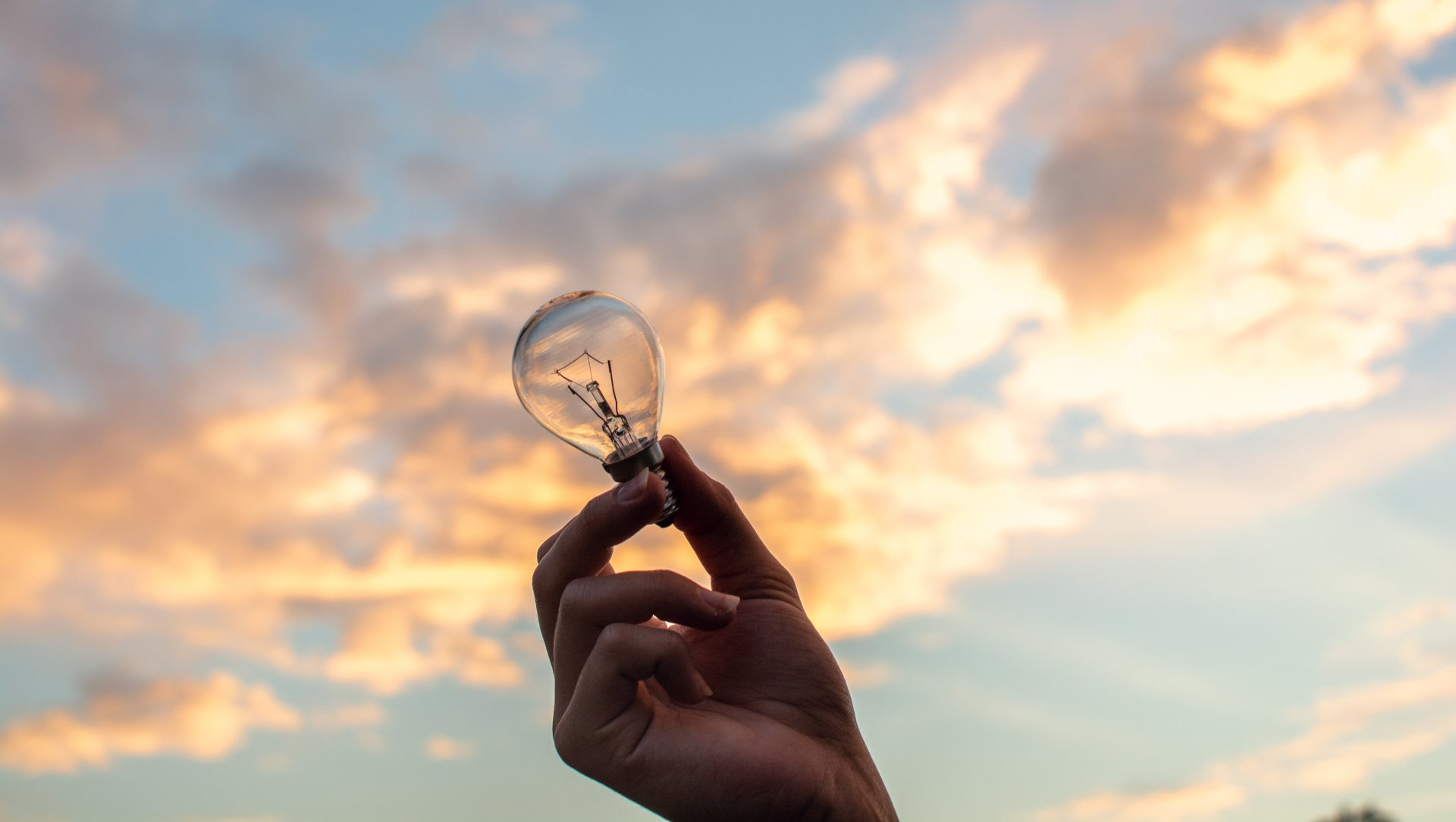 A person is holding a light bulb in their hand in front of a cloudy sky.
