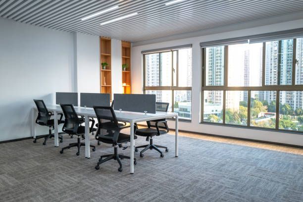 Office space with desks, chairs, and computers near large windows overlooking cityscape.