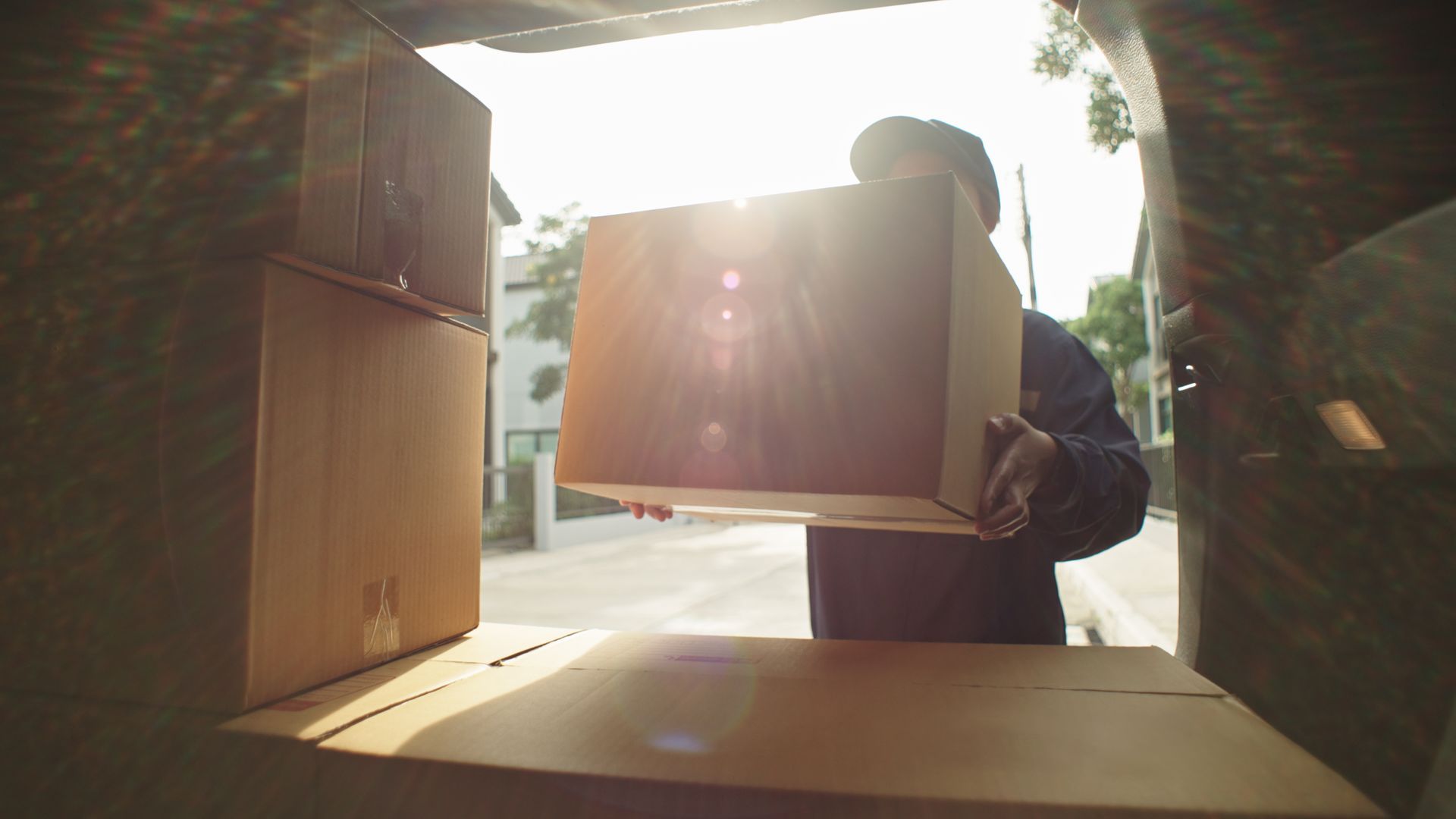 Person loading large cardboard box into the back of a vehicle. Bright sunlight filters through.