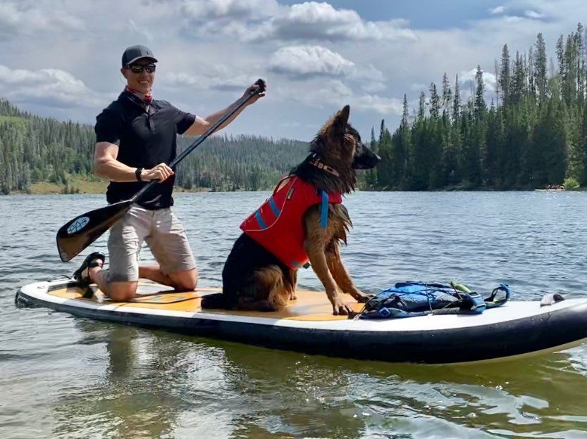 A man and a dog are on a paddle board in the water . the dog is wearing a life jacket.