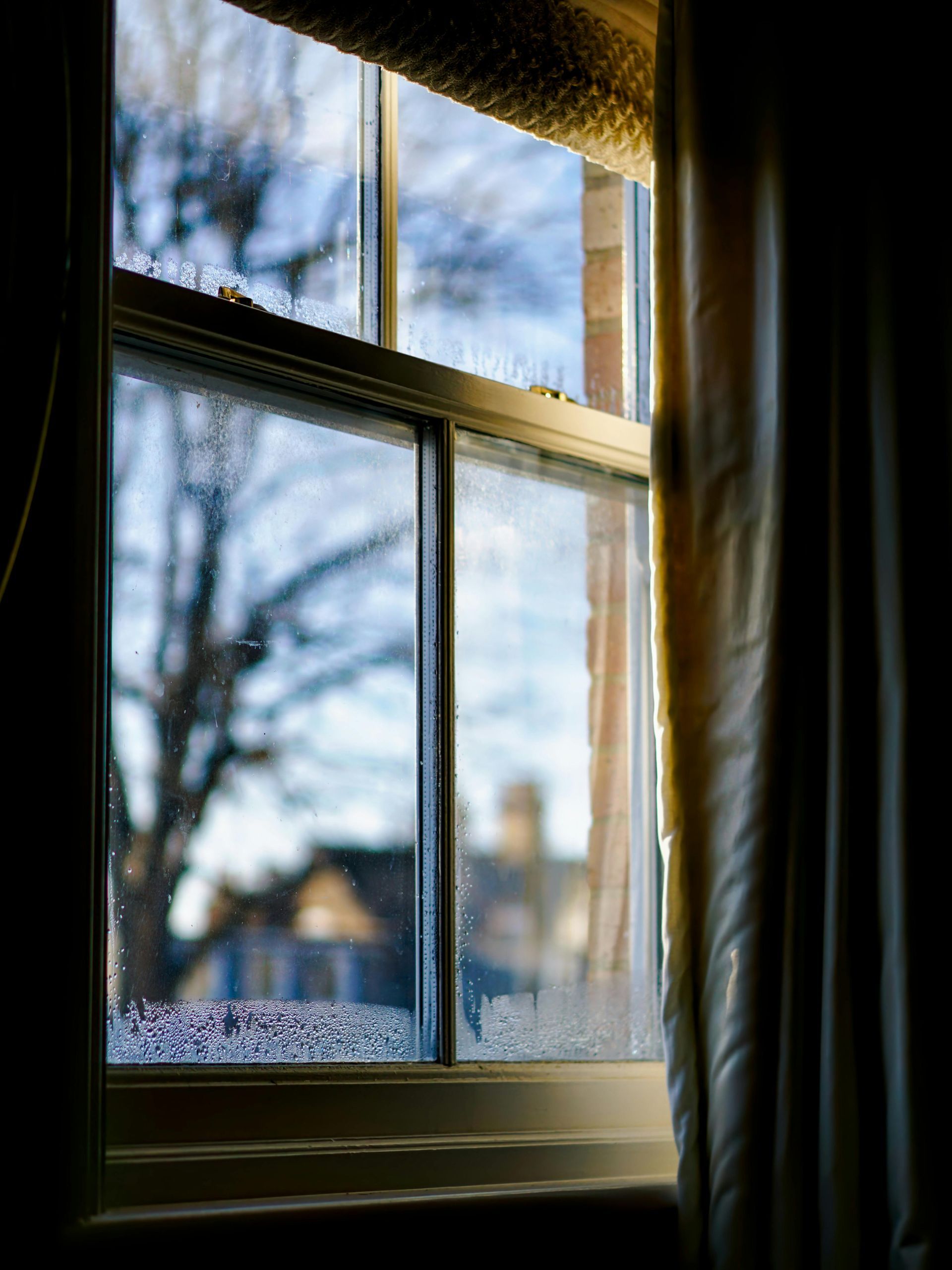 Window with condensation, partially open, showing blurry trees, houses, and sky. Beige curtain to the right.