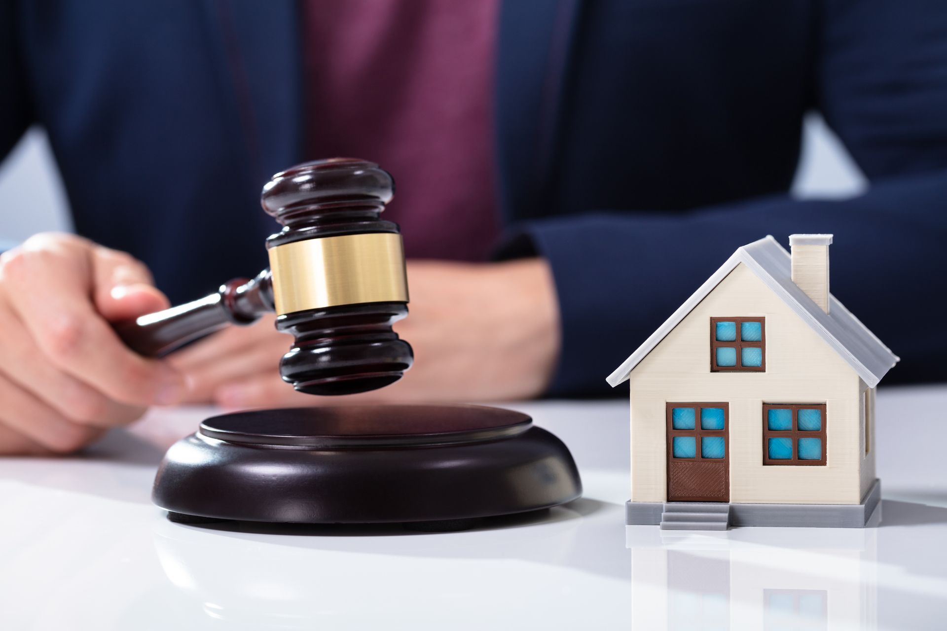 Real estate attorney is striking wooden gavel beside a small house model on a white table surface.