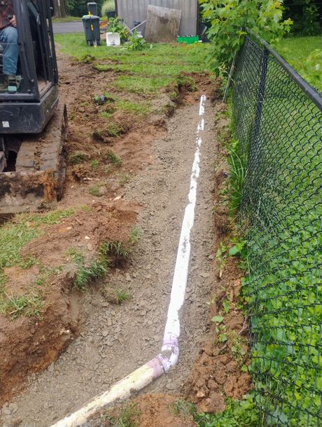 Trench with white pipe being installed next to a black fence; small excavator present.