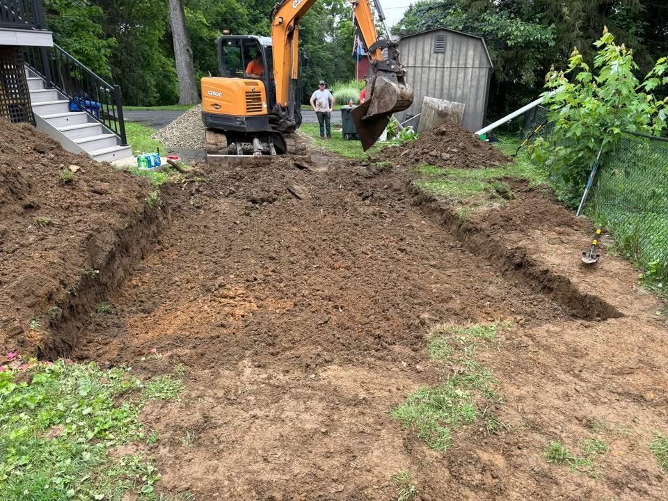 An excavator digs in a backyard, creating a rectangular trench. A person watches nearby.