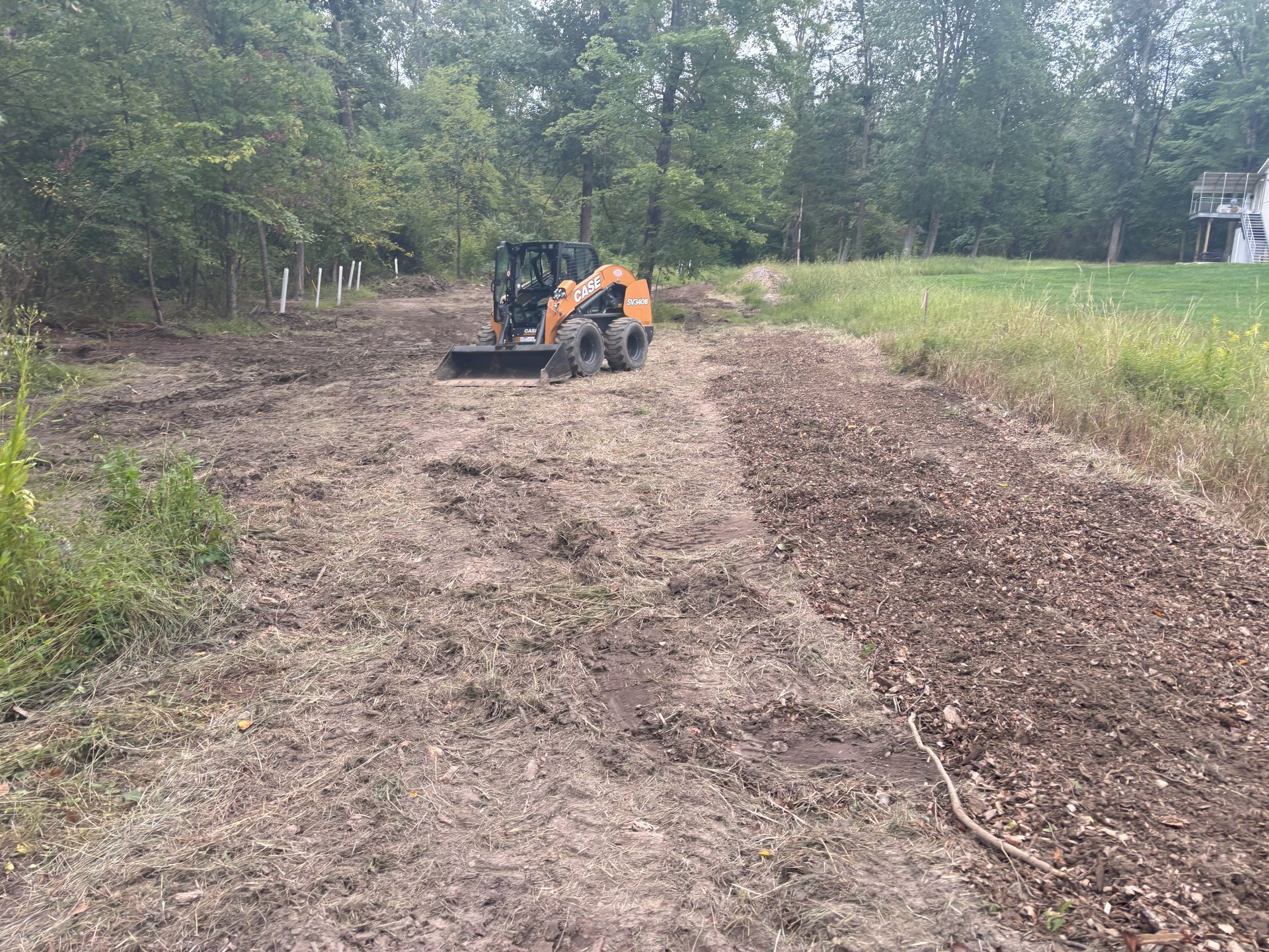 Skid steer grading dirt path near a tree line and grassy field.