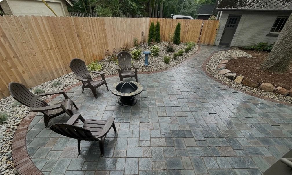 Patio with fire pit, chairs, and landscaping. Brown wooden fence, brick pavers, and gravel surround.