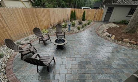Patio with fire pit, chairs, and landscaping. Brown wooden fence, brick pavers, and gravel surround.