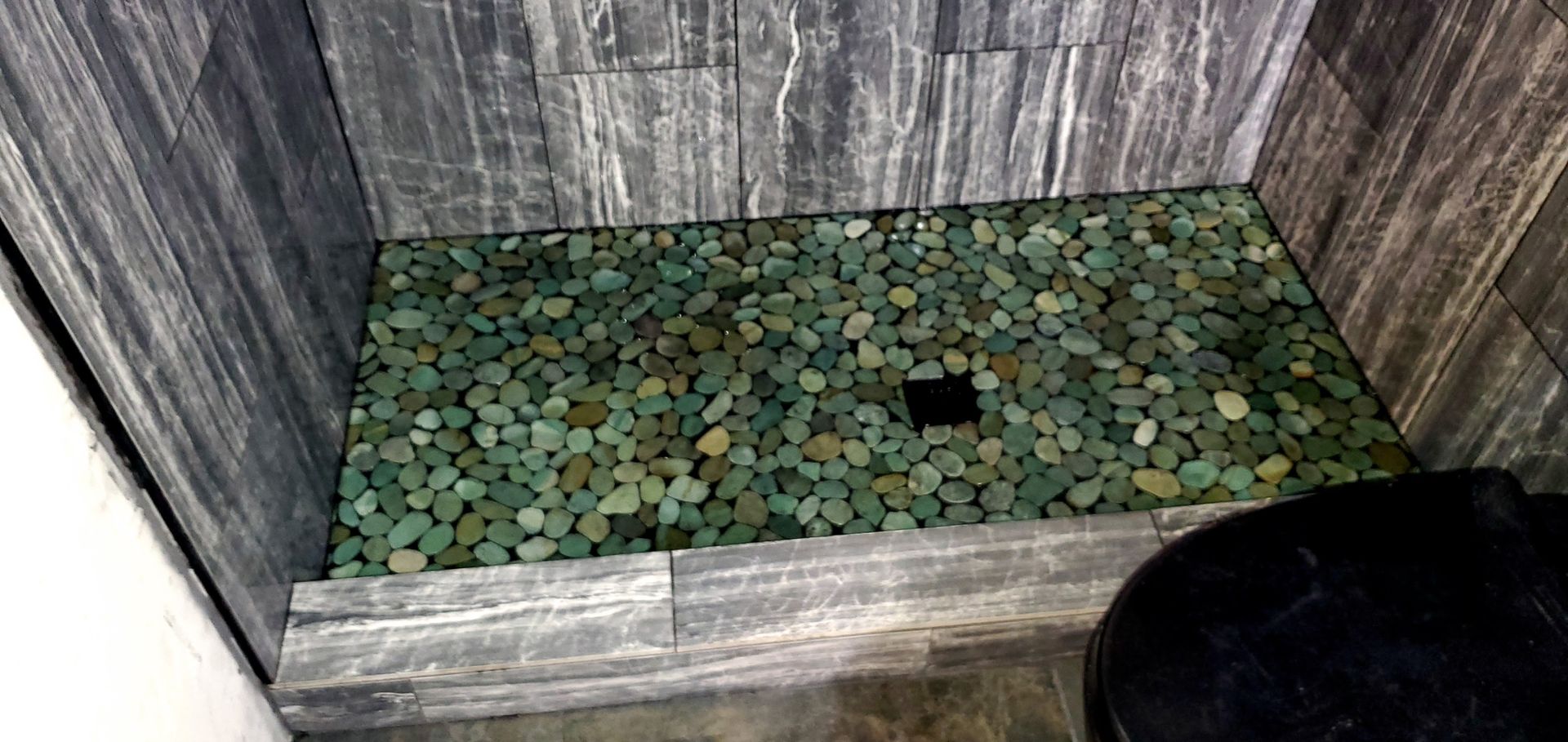 A shower stall with a green tile floor and a black toilet.