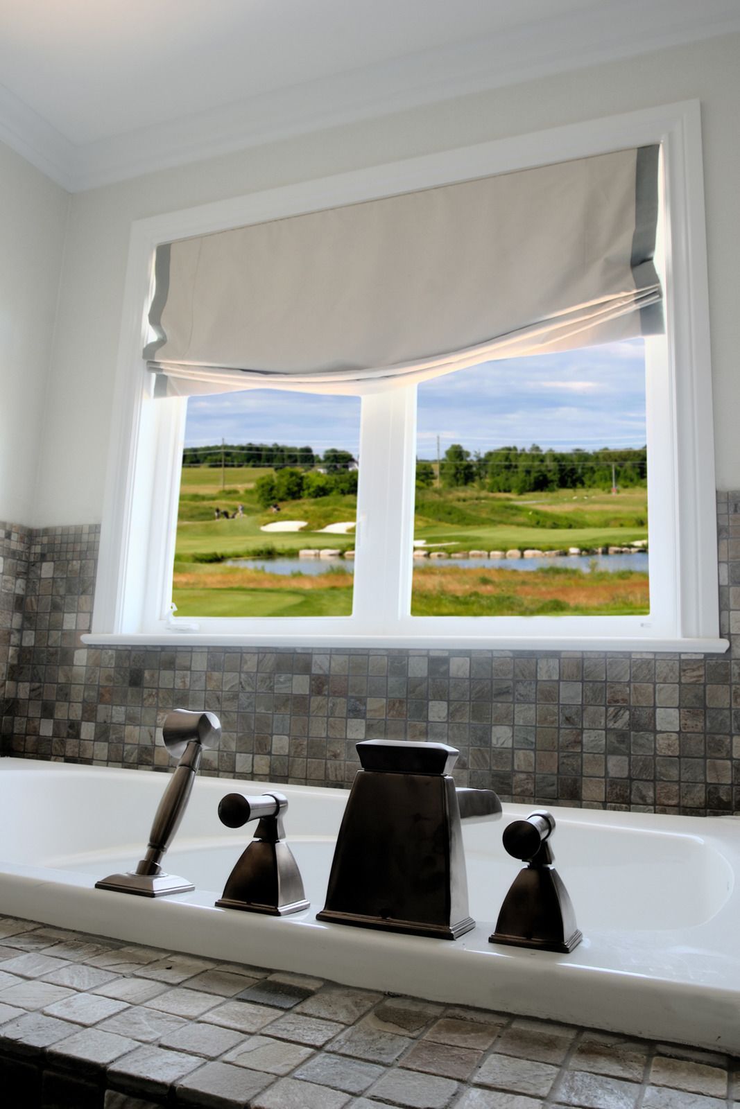 A bathtub with bronze fixtures in front of a window overlooking a golf course. The window has a Roman shade.