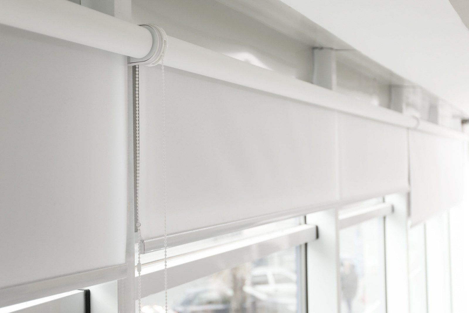 White roller shades partially covering a window. The shades have a metal chain and are mounted on a white track.
