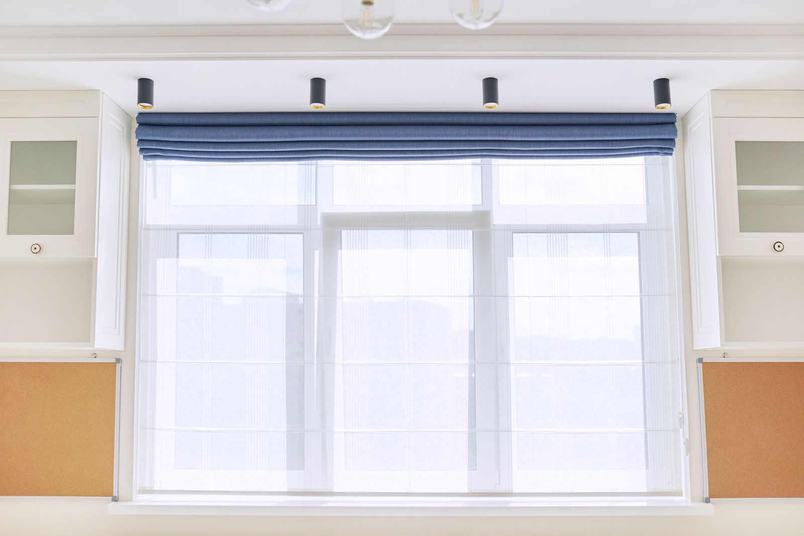 A bright window with a blue shade, sheer curtains, and overhead lighting in a room with cabinets and wood panels.