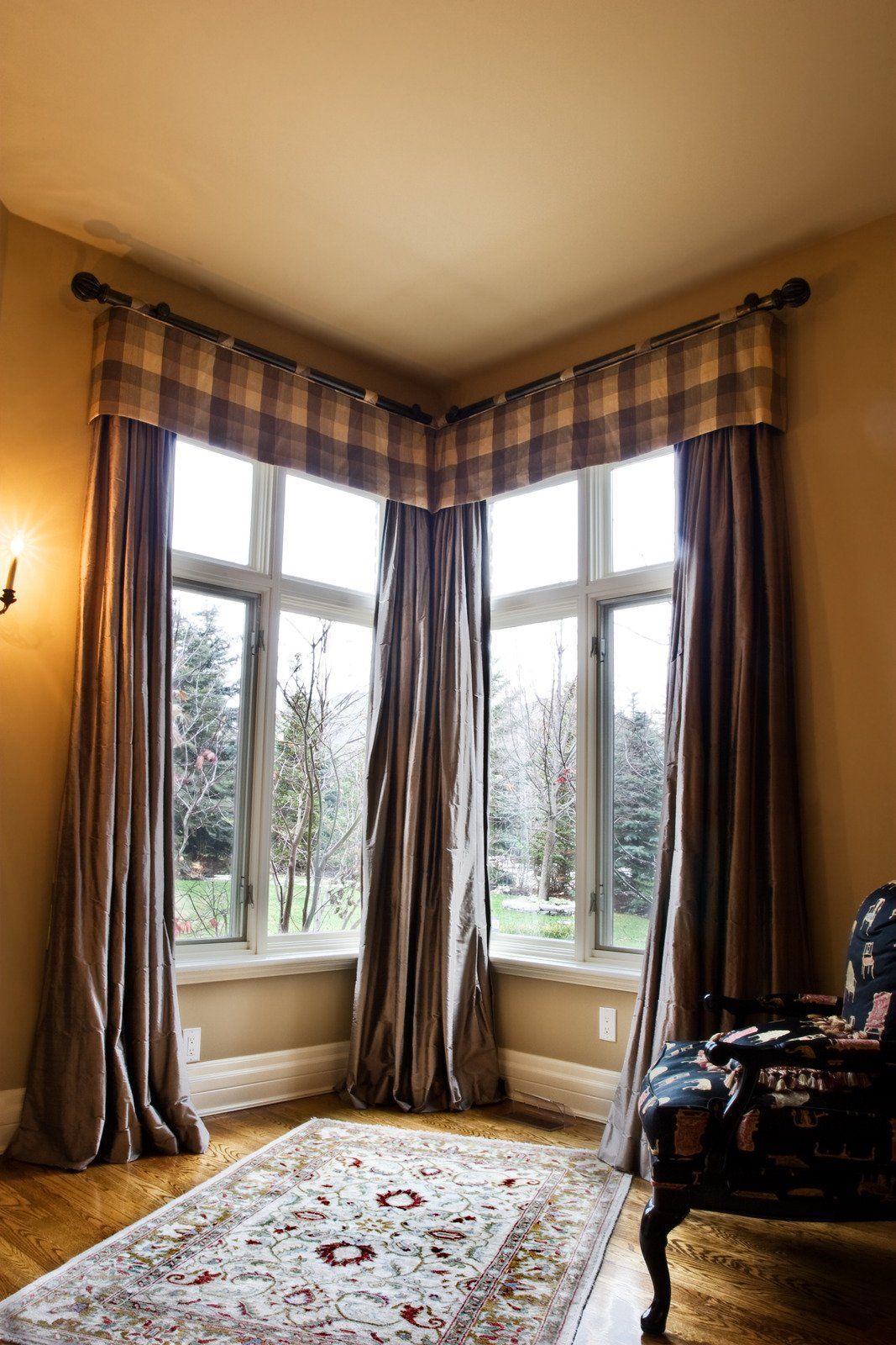 A corner window with a checkered valance and brown curtains. An oriental rug sits on the hardwood floor.
