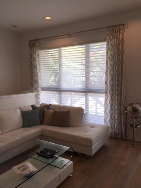 Living room with white sectional sofa, glass coffee table, window with blinds and patterned curtains, and hardwood floor.