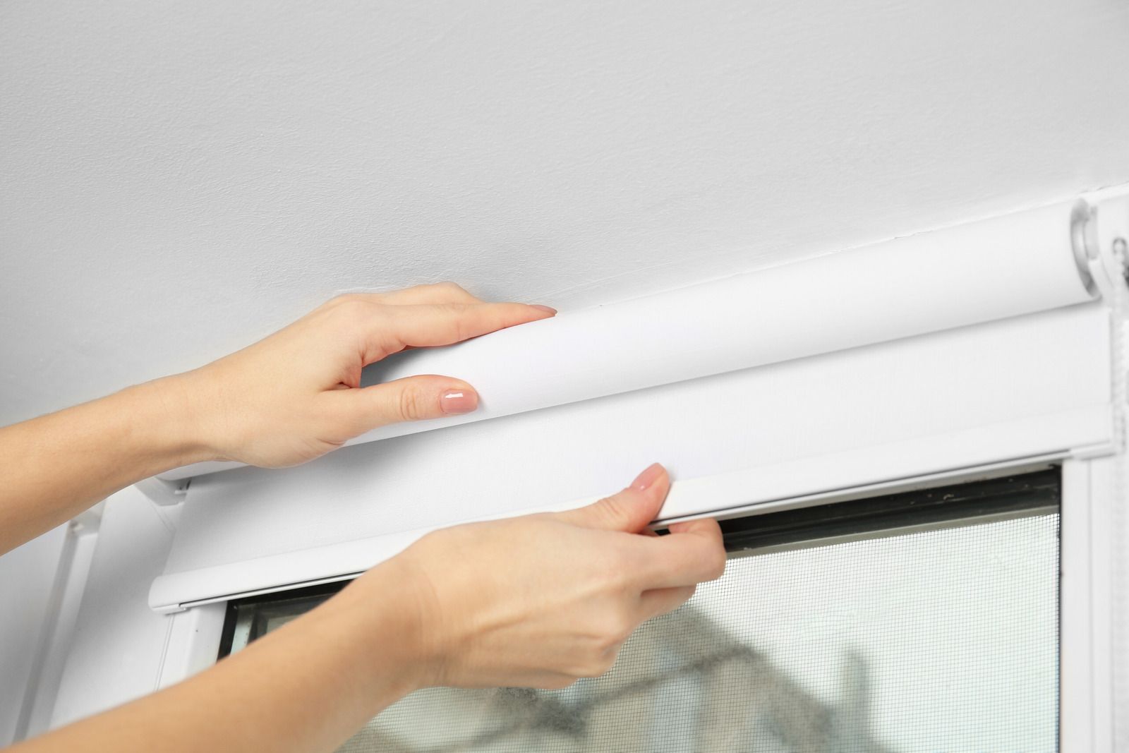 Hands adjusting a white roller blind in front of a window.
