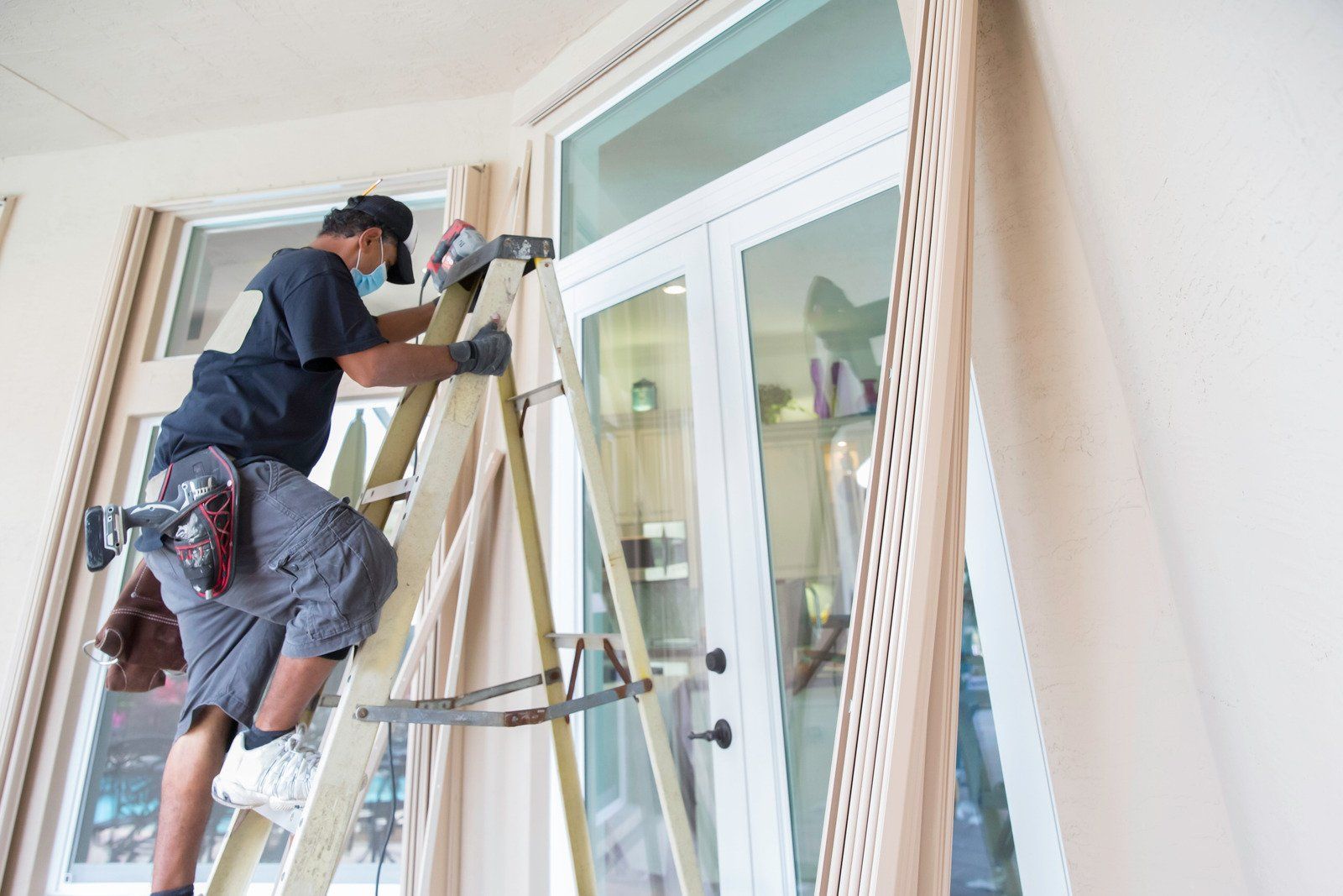 Man on a ladder cleaning a glass door in a room with beige curtains. He wears a cap, mask, shorts, and gloves.