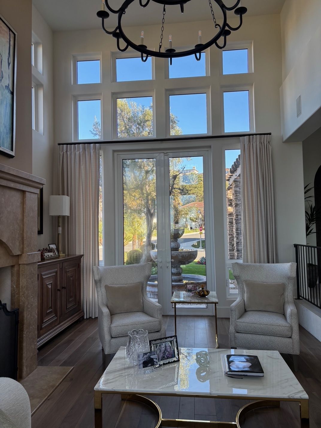 Living room with tall windows, a chandelier, and seating area; neutral color scheme with wood flooring, and outdoor view.