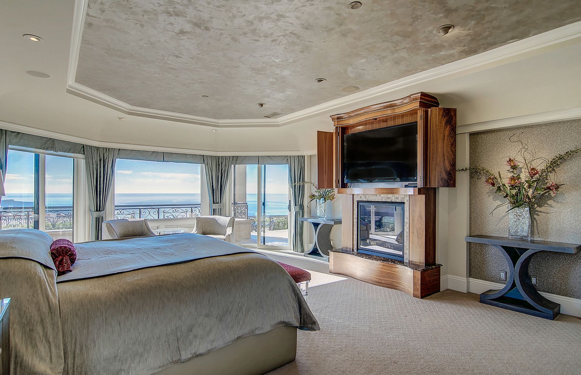 Spacious bedroom with large windows overlooking a cityscape. A large bed, fireplace, and seating area are visible.