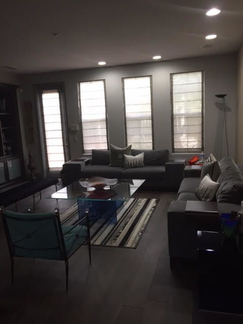 Modern living room with gray sofas, a glass coffee table, and large windows with shades. Dark flooring and walls.