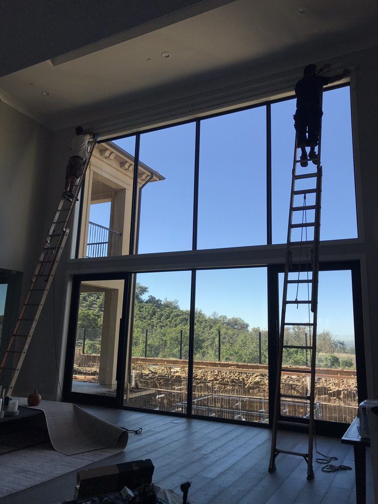 Two workers installing window film on tall, floor-to-ceiling windows. One is on a ladder, the other on the ground.