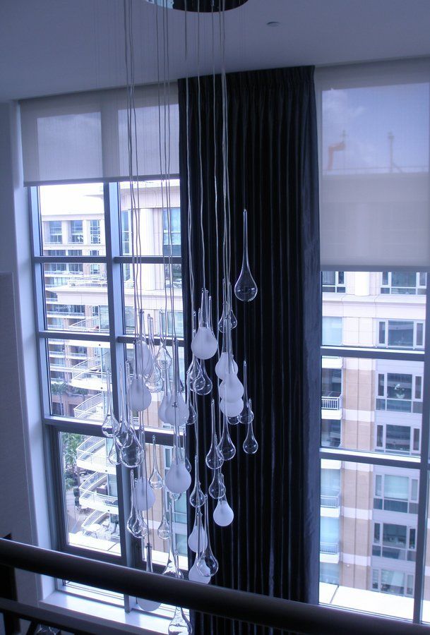 A tall, modern chandelier hangs in front of a large window with black drap
