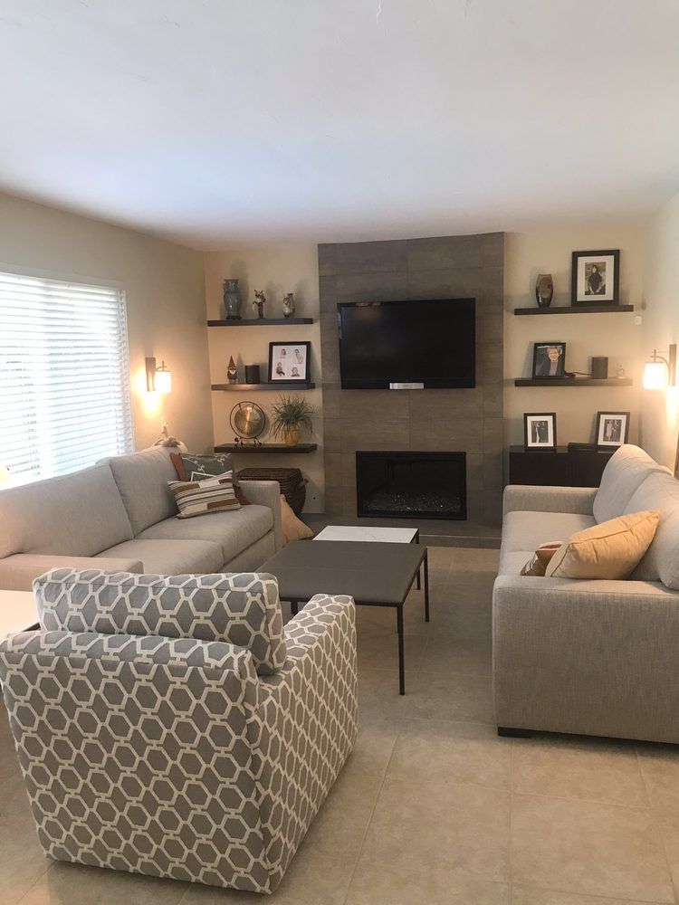 Living room with gray couches, patterned armchair, fireplace with TV, and decorative shelves. Beige walls and tile floor.