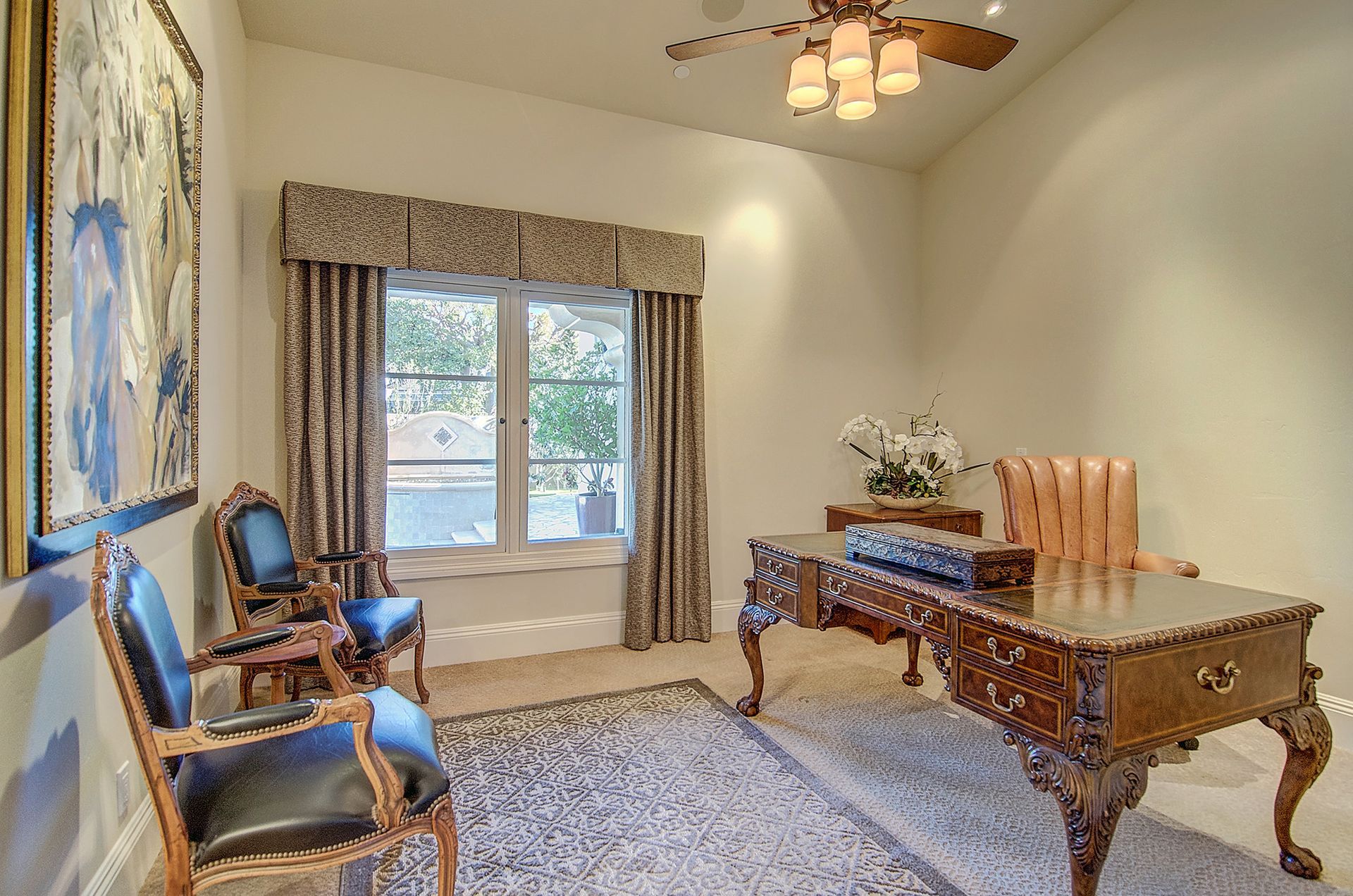 An ornate home office featuring a large wooden desk, two blue chairs, a tan leather desk chair, and a window with curtains.