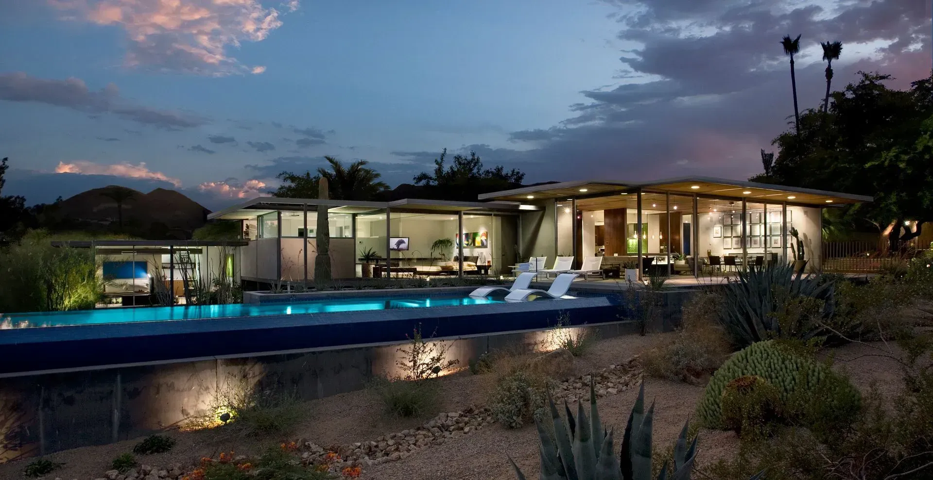 Modern home with a pool at dusk; glass walls, white furniture, and desert landscaping.
