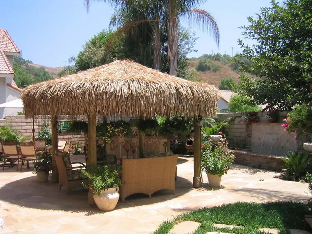 Thatched-roof gazebo with seating in a backyard patio with potted plants and a fountain.