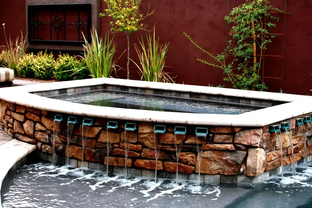 Stone-walled rectangular water feature with spouts, water flowing into a pool, set against a red stucco wall