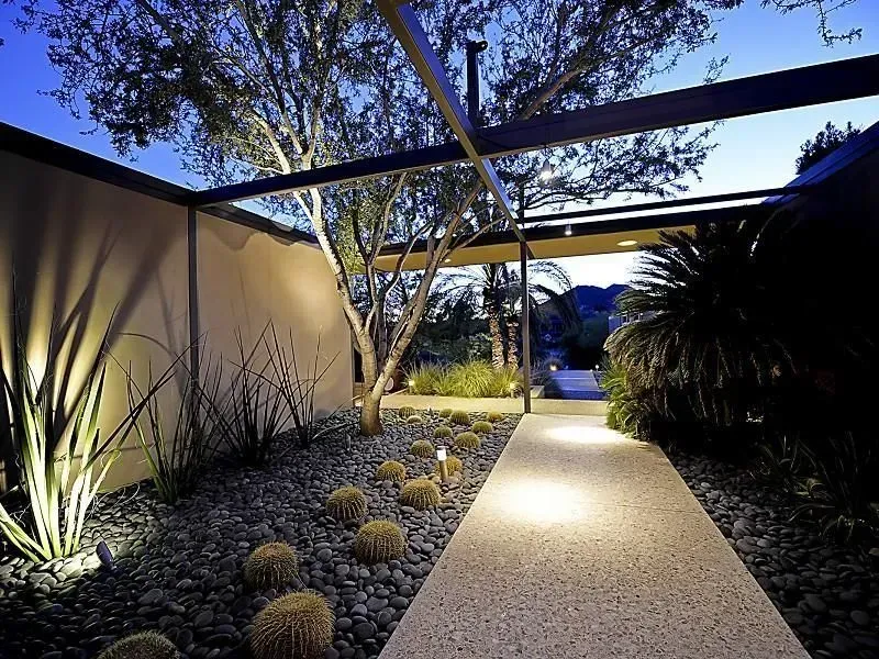 Pathway with plants and spotlights, leading to a landscape under a metal structure.