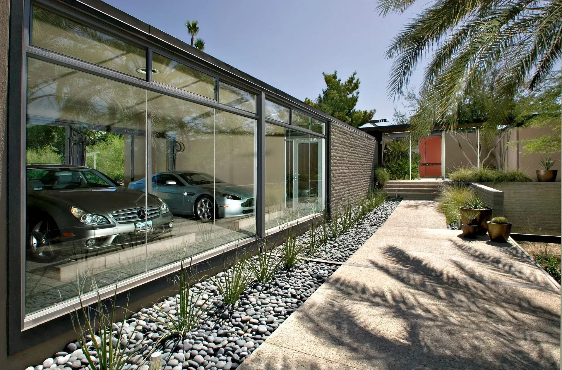 Glass-walled garage with two cars parked inside, next to a walkway with landscaping under a bright blue sky.