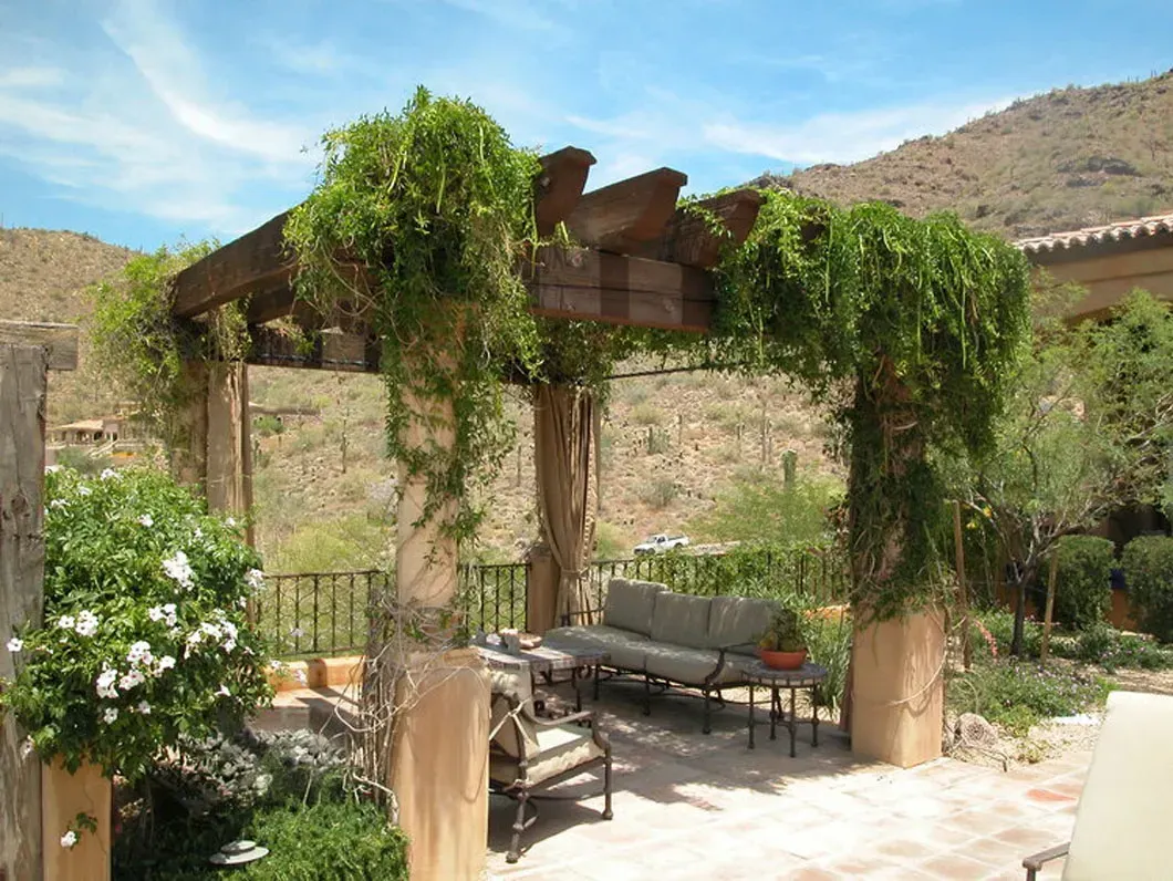 Wooden pergola covered in green vines over a patio with outdoor seating, desert mountains in the background.