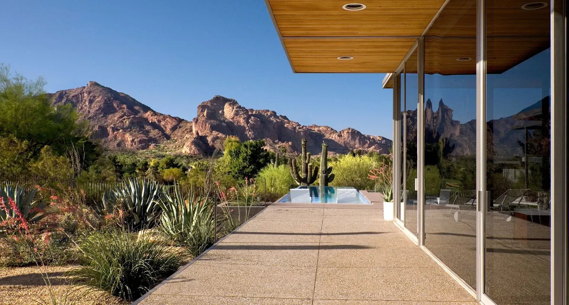 Long, modern home with floor-to-ceiling windows and a pool, overlooking desert landscape and mountains under a blue sky.