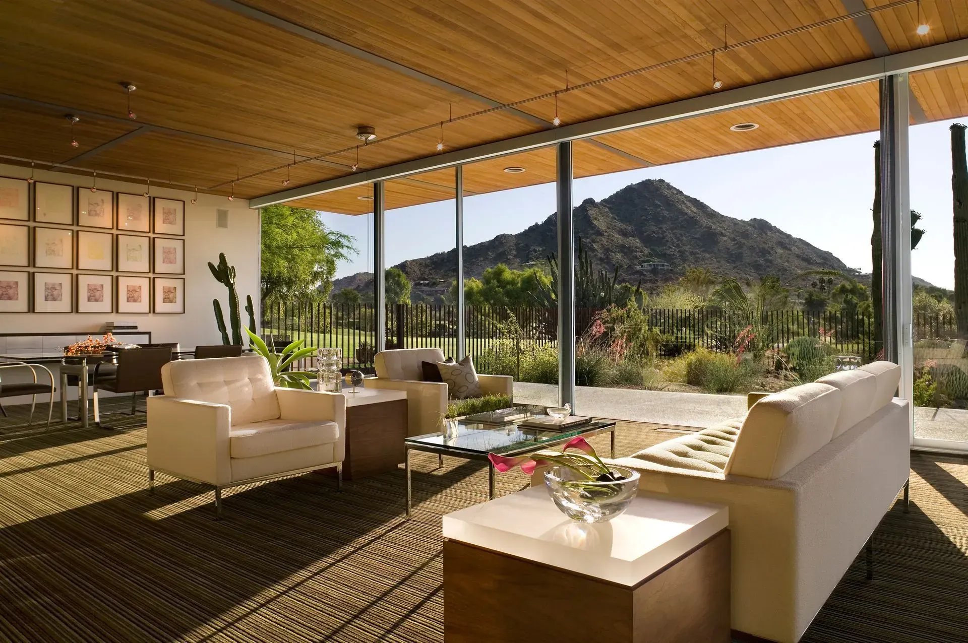 Living room with large windows overlooking a mountain and desert landscape. Light-colored furniture and wood ceiling.