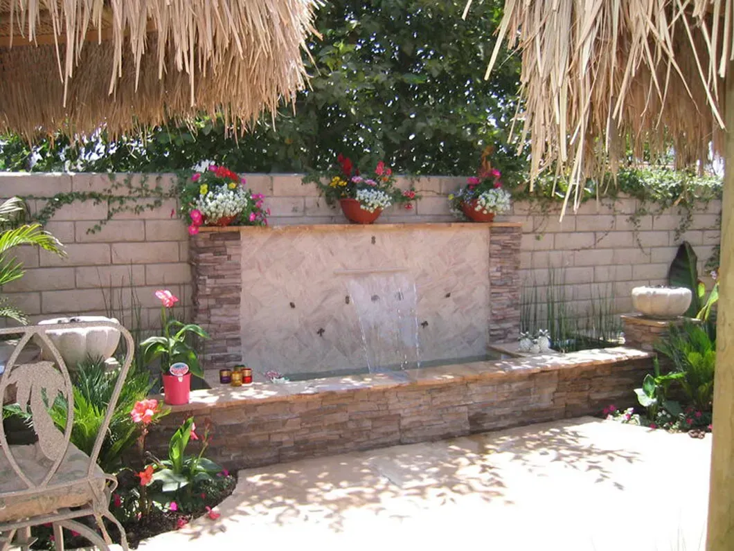 Backyard water feature with stone wall, flowers, and thatch-roof shade.